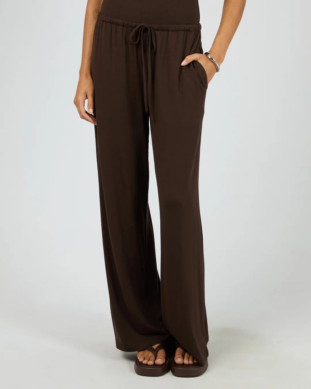 A person wearing Dara dark brown wide-leg drawstring pants with side pockets, paired with matching dark brown sandals, shown against a light gray studio background.